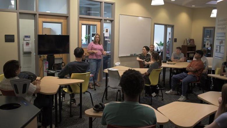 Person stands at the front of a room, talking to other people sitting at tables. 